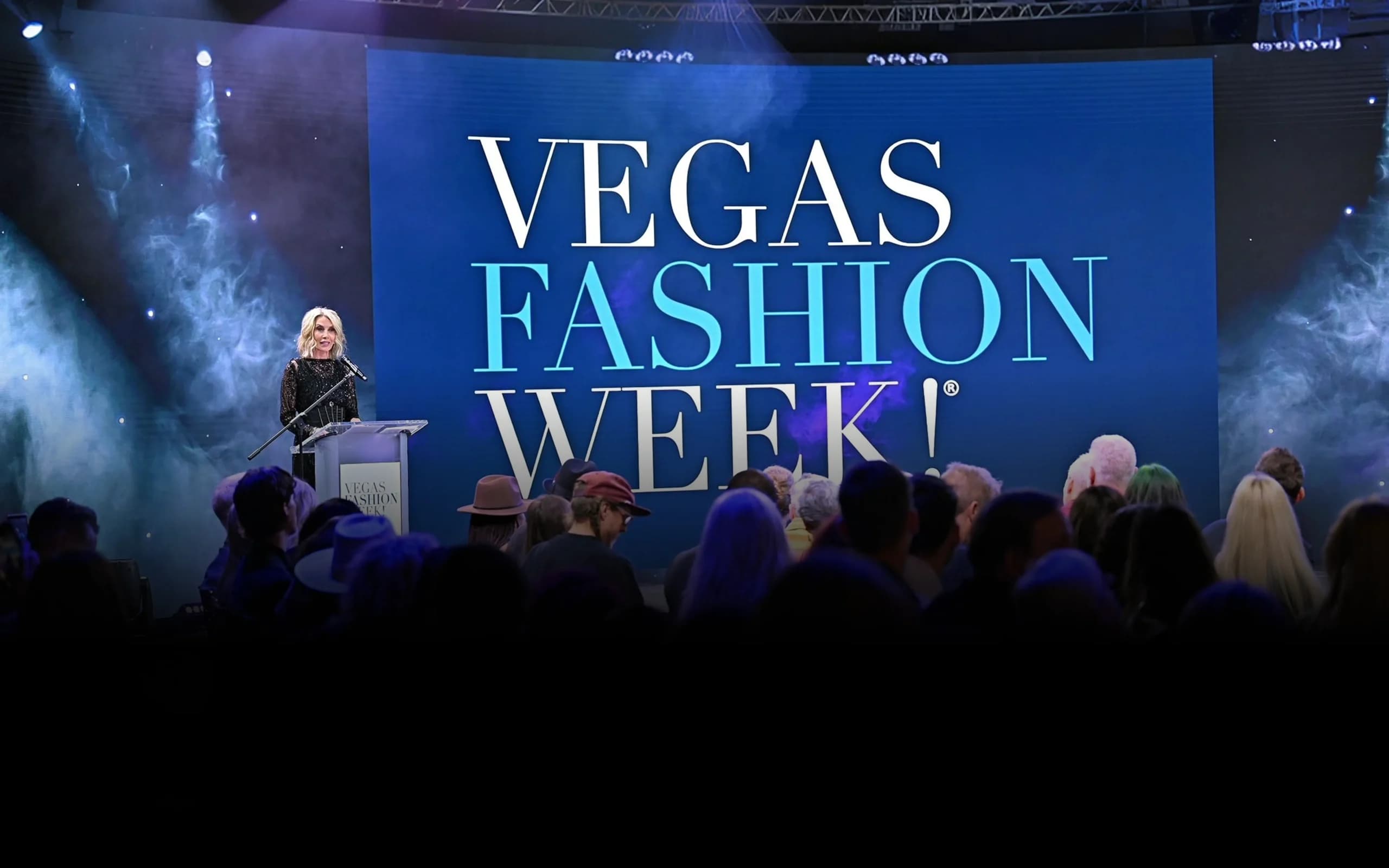 A speaker addressing the audience at Vegas Fashion Week, with the event's name shown on a stage screen behind them