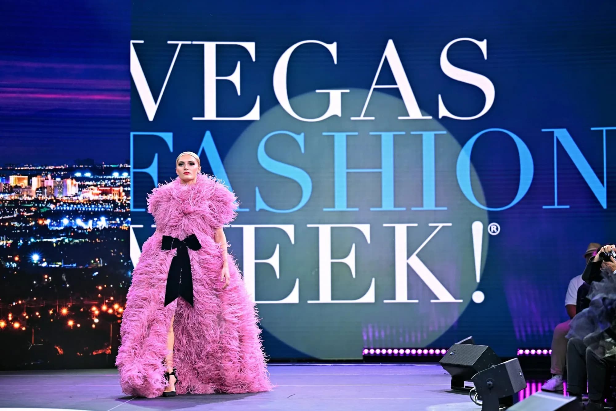 A model presents a designer's look on the runway at Vegas Fashion Week®, with the event's name on the stage screen behind